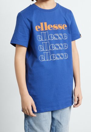 Dziecko noszące niebieską koszulkę z nadrukiem "ellesse" czterokrotnie w kolorze pomarańczowym i białym, połączoną z jasnoniebieskimi dżinsami.
