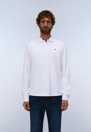 Homme aux cheveux bouclés et à la barbe, portant un polo blanc à manches longues et un jean bleu foncé, debout devant un fond clair et uni.