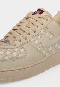 Μπεζ αθλητικό παπούτσι Nike με υφασμάτινες λεπτομέρειες στο πάνελ, χοντρά κορδόνια και κόκκινη ετικέτα στη γλώσσα με το λογότυπο Nike Air.
