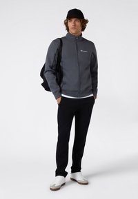 Grijze sportjack met ritssluiting en hoge kraag, zwarte joggingbroek en witte sneakers. Het model draagt een zwarte rugzak.