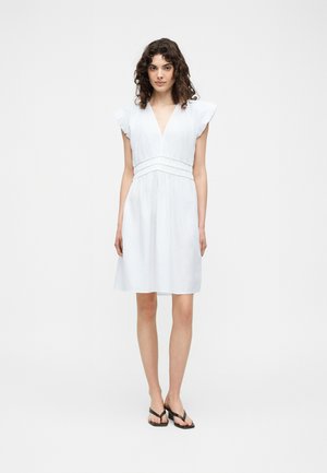 Γυναίκα που στέκεται, φοράει ένα λευκό φόρεμα μέχρι τα γόνατα με βολάν στα μανίκια, βαθύ V-λαιμόκοψη, σούρες στη μέση και μαύρα σανδάλια με τακούνι.