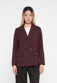 Bordeaux dobbeltradet blazer i tekstureret stof, med to forlommer og sorte knapper, parret med en hvid top og mørke bukser.