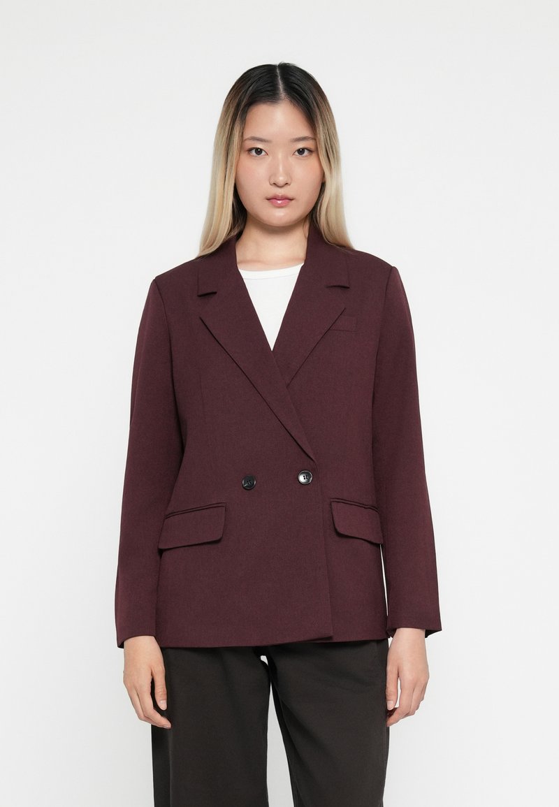 Bordeaux dobbeltradet blazer i tekstureret stof, med to forlommer og sorte knapper, parret med en hvid top og mørke bukser.