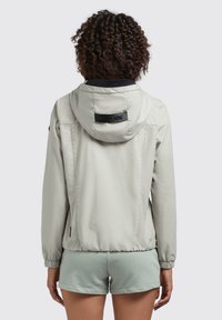 Lichtgrijze zip-up jack met een capuchon, elastische manchetten en een zwart logoaccent op de achterkant. Gladde, matte textuur; ontworpen voor comfort.