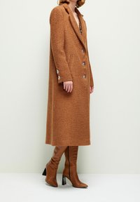 Femme portant un long manteau marron texturé avec de gros boutons, debout en bottes en cuir marron à talons montant jusqu'aux genoux, sur un fond uni.