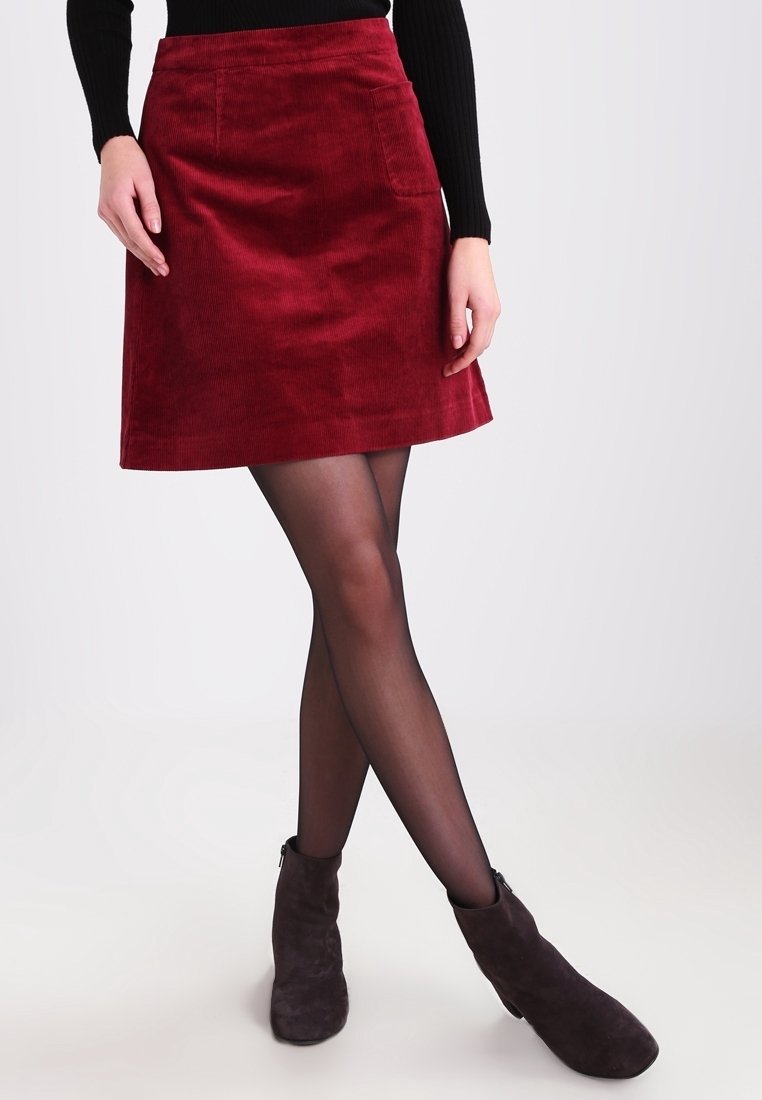 Falda A-line de pana roja con bolsillos de parche, textura suave y dobladillo por encima de la rodilla, combinada con botas de tobillo oscuras y medias negras.