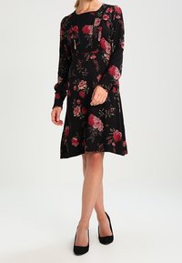 Vestido negro con flores que presenta mangas largas, un corpiño ajustado y una falda acampanada; adornado con un patrón de rosas rojas y rosas.