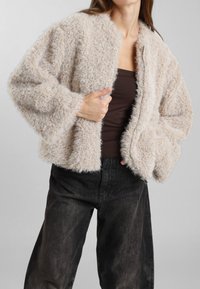 Giacca corta fuzzy beige con maniche oversize e scollatura rotonda, dotata di zip frontale e realizzata in un tessuto morbido e testurizzato.