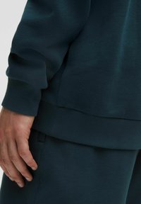 Tyrkysový sveter s rebrovanými manžetami a lemom, vyrobený z jemného materiálu. Obsahuje malé, embosované logo blízko lemu. Ruka odpočíva na boku.