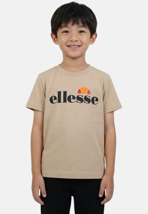 Jeune garçon souriant, portant un t-shirt beige avec le logo "ellesse" noir et orange, et un pantalon noir, debout devant un fond uni.