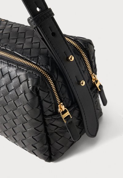 Μαύρη υφαντή δερμάτινη τσάντα με δύο χρυσά φερμουάρ και ρυθμιζόμενο λουράκι με χρυσά τρουκς σε λευκό φόντο.