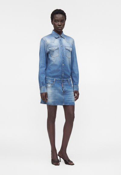 Γυναίκα με ξεθωριασμένο μπλε τζιν πουκαμισοφόρεμα με τσέπες στο στήθος και λεπτομέρειες με κουμπιά, φοράει μαύρες ανοιχτές ψηλοτάκουνες γόβες, στεκόμενη μπροστά σε λευκό φόντο.