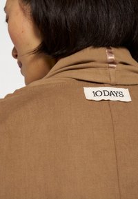 Camicia in lino marrone con un'etichetta posteriore su un patch crema testurizzato con la scritta "10DAYS" in nero, che mette in evidenza una leggera lucentezza nel tessuto.
