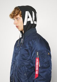 Mornarska bomber jakna s sjajnom površinom, rebrastim manžetama i džepom na patentni zatvarač. Priključena crna kapuljača sadrži izrazite bijele natpise i vezice.