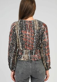 Femme aux longs cheveux bruns portant une blouse florale texturée multicolore avec taille smockée et fermeture à boutons au dos, associée à un jean en denim gris.