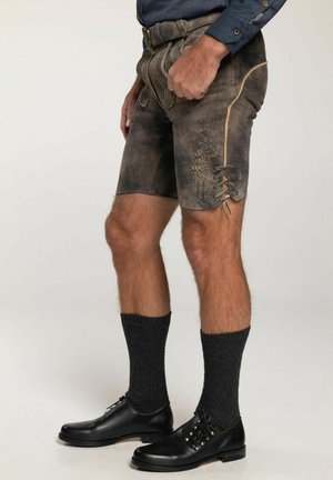 Dunkle Lederhosen mit dekorativer Stickerei, seitlicher Schnürung und taillierter Passform, kombiniert mit dunklen Strumpfhosen und schwarzen Schuhen.