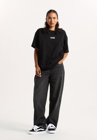 Camiseta de algodón negra con el logo de Vans, corte holgado, combinada con unos vaqueros de pierna ancha en gris oscuro y zapatillas en blanco y negro con cordones blancos.