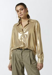 Zlatna metalik košulja na dugih rukava s ovratnikom, nosi se uz karirane hlače visokog struka i crni remen.