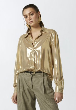 Camicia dorata metallizzata con colletto e maniche lunghe, indossata con pantaloni a quadri a vita alta e una cintura nera.
