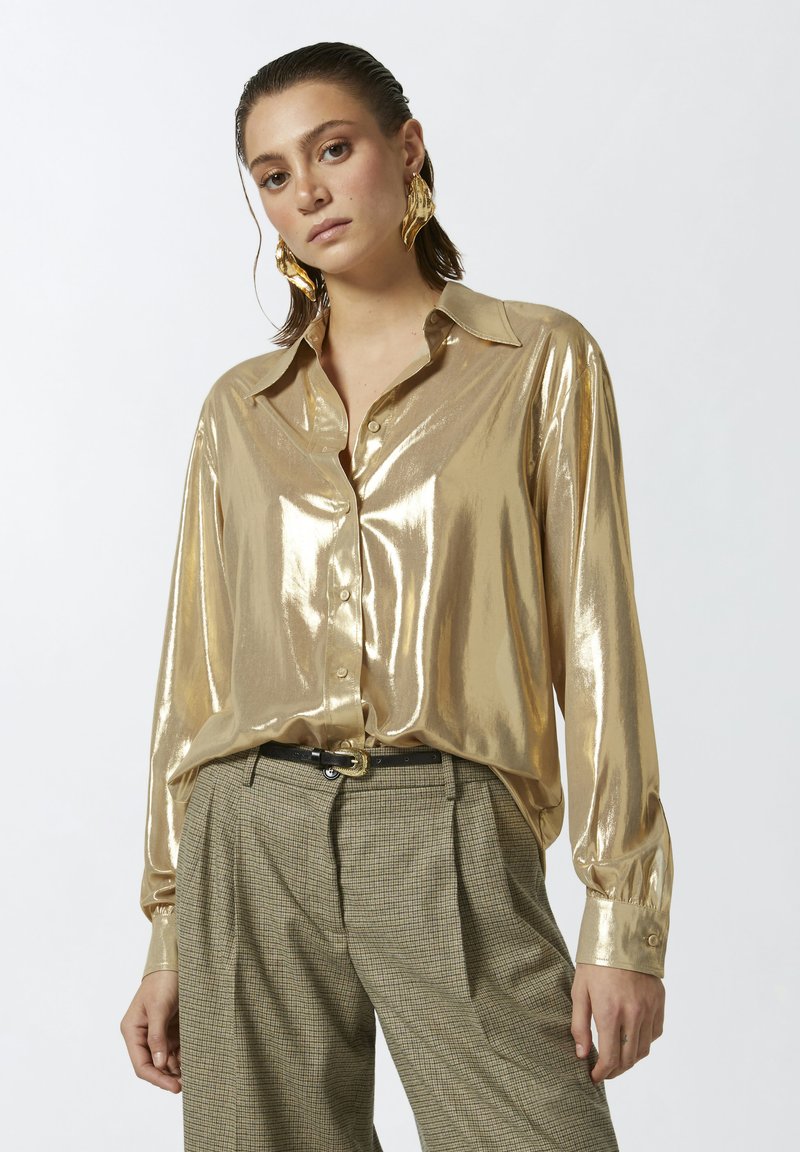 Zlatna metalik košulja na dugih rukava s ovratnikom, nosi se uz karirane hlače visokog struka i crni remen.