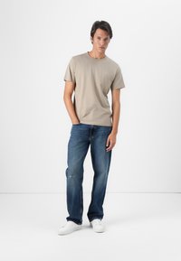 Light beige cotton t-shirt with short sleeves, paired with blue denim jeans. Model stands with hands in pockets, wearing white sneakers.