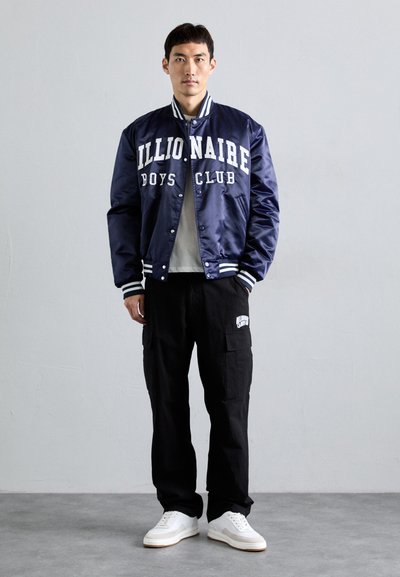 Mężczyzna w granatowej satynowej kurtce varsity z napisem "Billionaire Boys Club", czarnych spodniach cargo i białych sneakersach, stojący przy gładkiej ścianie.