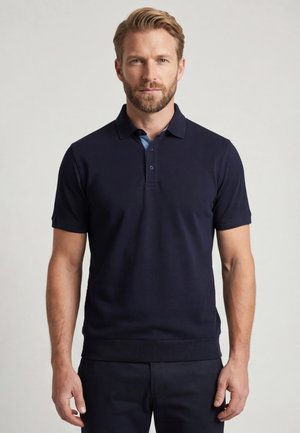 Baarddragende man met lichtbruin haar, dragend een donkerblauw poloshirt met korte mouwen en bijpassende donkere broek, staand tegen een effen achtergrond.