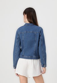 Mujer con cabello largo castaño, vistiendo una chaqueta de mezclilla azul y pantalones cortos blancos, de pie frente a una pared lisa de color gris claro.