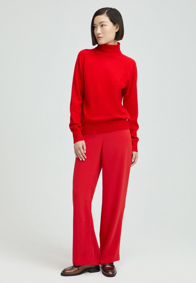 Roter Pullover mit hohem Kragen und langen Ärmeln, kombiniert mit passenden roten weit geschnittenen Hosen; das Modell trägt braune Schuhe, schlichtes Design, glatte Textur.