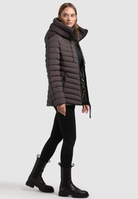 Dunkelbraune gepolsterte Jacke mit Kapuze, vertikal gestepptem Design, mit Reißverschlüssen versehenen Taschen und Kordelzug-Details. Getragen über schwarzen Leggings und Stiefeln.