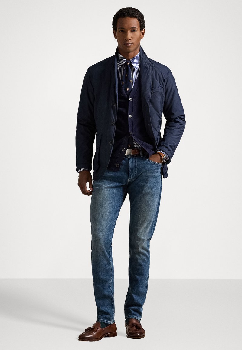 Navy quilted jacket over a dark vest, striped shirt, and tie. Light blue jeans and brown loafers complete the look. Simple, tailored design.