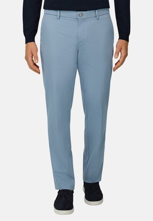 Mann trägt hellblaue, maßgeschneiderte Hose, schwarze Slipper und ein marineblaues Langarmshirt, steht mit entspannten Händen an den Seiten.