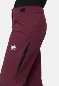 Pantalon softshell grenat avec poche zippée sur la cuisse, écusson logo, patte de réglage à la taille, porté avec un haut noir.