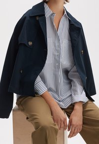 Marineblauer Trenchcoat mit großen Knöpfen über einem weiß gestreiften Hemd. Hellbraune Hosen. Glattes Material und lockere Passform. Minimalistisches Design.