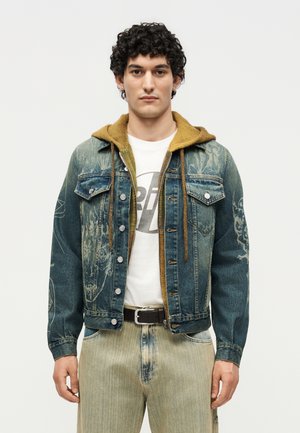 Młody mężczyzna w niebieskiej dżinsowej kurtce z brązowym kapturem, białej koszulce z nadrukiem, beżowych spodniach i czarnym pasku, stojący na jednolitym tle.