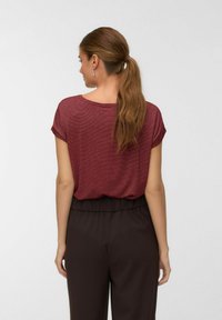 Kvinde med lang hestehale iført en kortærmet bordeaux top og løse sorte bukser, stående med ryggen mod en ensfarvet baggrund.