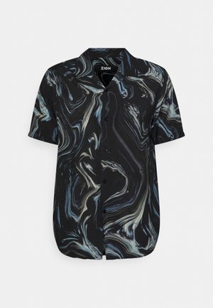 Homme portant une chemise noire à manches courtes avec des tourbillons bleus et blancs, un pantalon gris et des sandales en cuir noir tressées, se tenant avec une main dans la poche.