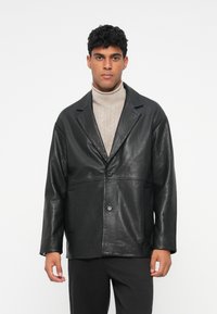 Giacca di pelle nera con una finitura testurizzata, chiusura a un pulsante e due tasche anteriori, indossata sopra un dolcevita beige chiaro.