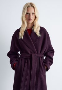 Plüschiger, bordeauxfarbener Wickelmantel mit einem Bindegürtel, weiten Ärmeln und Seitentaschen. Die gewebte Textur verleiht Tiefe und Wärme.