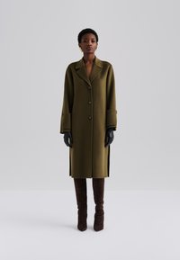 Abrigo de lana verde oliva con cuello muesca, botones delanteros, puños anchos y aberturas laterales, combinado con guantes negros y botas marrones hasta el muslo.