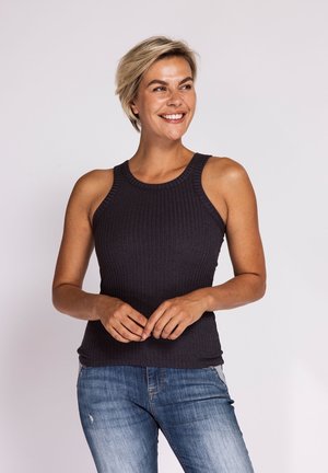 Lächelnde Frau mit kurzen blonden Haaren, die ein ärmelloses schwarzes, geripptes Top und blaue Jeans trägt, steht vor einem schlichten hellen Hintergrund.