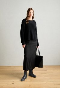 Must kulunud kahekihiline kampsun musta pikkade seeliku peal, hallide nööridega saapadega ja musta käeketiga. Puitpõranda taust.