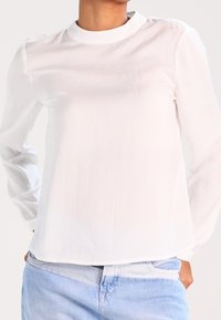 Top bianco a maniche lunghe con colletto rotondo, texture liscia, spalle basse e orlo dritto, abbinato a jeans azzurri chiari.