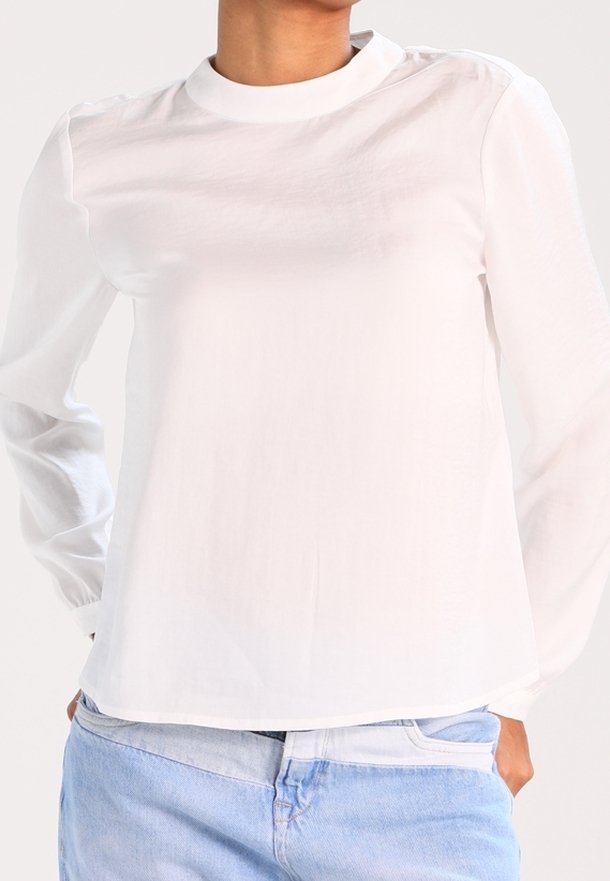 Top bianco a maniche lunghe con colletto rotondo, texture liscia, spalle basse e orlo dritto, abbinato a jeans azzurri chiari.