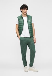 Zelená, prešívaná vesta bez rukávov cez biele tričko, v kombinácii s tmavozelenými teplákovými nohavicami. Oblečenie dopĺňajú krémovo biele tenisky.