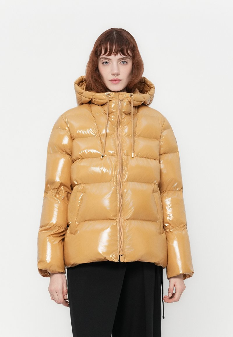 Donna che indossa un giubbotto puffer lucido color tan con cappuccio e zip frontale, in piedi contro uno sfondo bianco uniforme con le mani rilassate ai lati.
