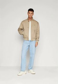 Jovem de pé com as mãos nos bolsos, vestindo uma jaqueta bomber bege, camisa branca, jeans azul-claro e sneakers brancos, contra um fundo cinza.