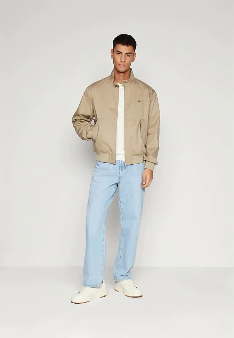 Jeune homme debout les mains dans les poches, portant une veste bomber beige, une chemise blanche, un jean bleu clair et des baskets blanches sur fond gris.