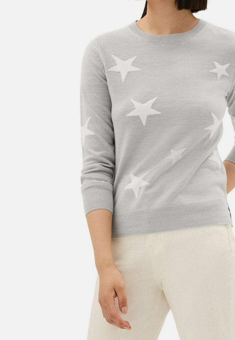 Femme portant un pull gris clair avec des motifs d'étoiles blanches et un pantalon blanc cassé, debout avec une main détendue le long du corps.