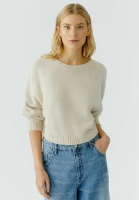 Licht beige pullovertrui met een geribbelde halslijn en mouwuiteinden, met een relaxte pasvorm en zachte textuur, gecombineerd met hooggeplaatste blauwe jeans.